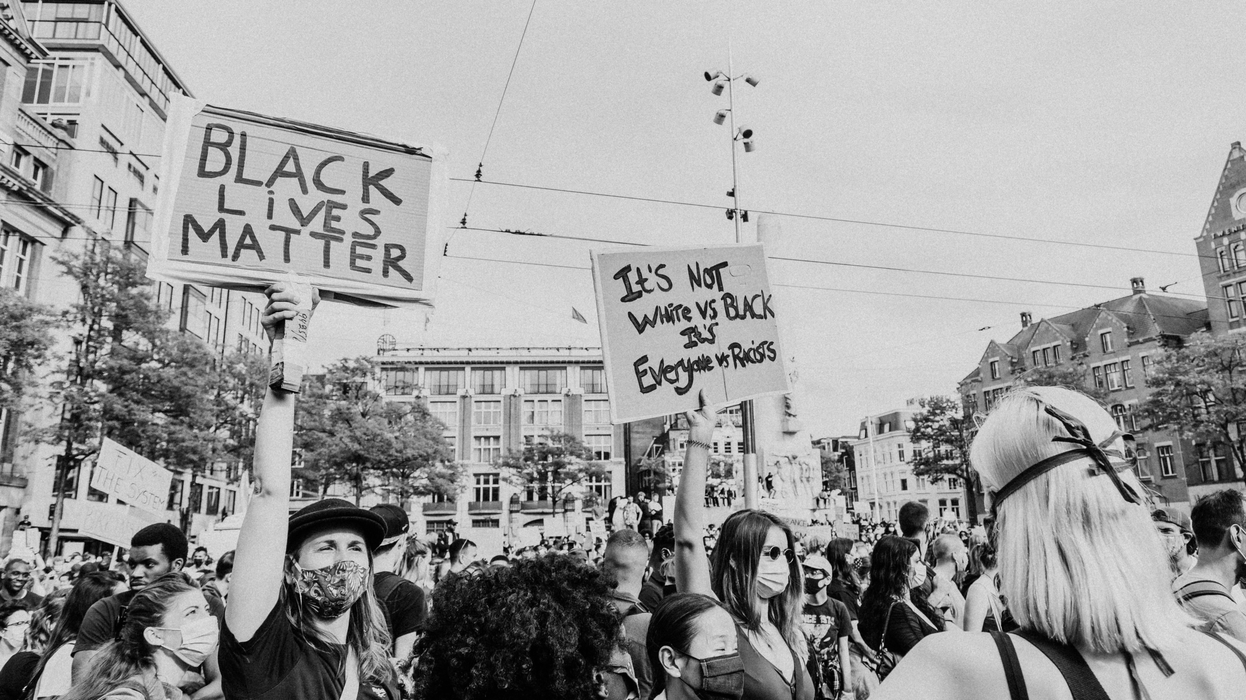 Black and white photo of a crowd protesting. Two signs are being held up, one reads "Black Lives Matter" and the other reads "It's not white vs black, it's everyone vs racists."